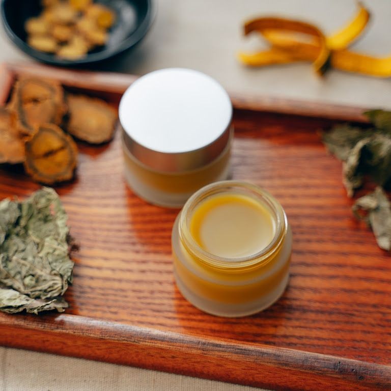 Two glass jars of herbal balm on a wooden tray with dried leaves and root slices.